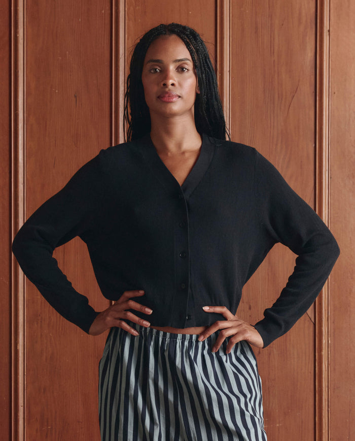 A person stands in front of a wooden panel wall, wearing THE GREAT. SP26 D1 The Shrunken V Neck Cardigan in black and striped pants, with hands on hips and a neutral expression.