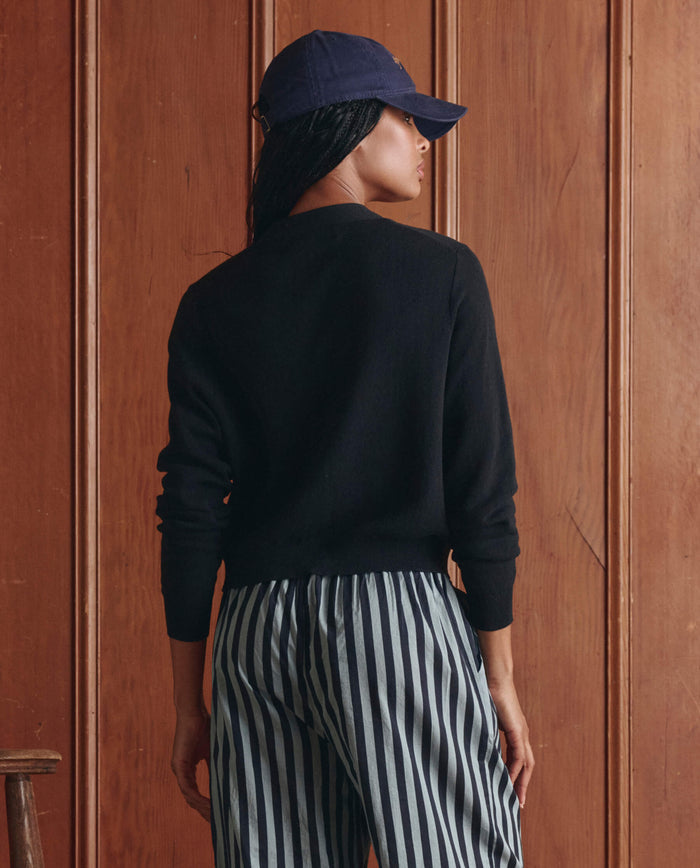 A person wearing a navy cap, black sweater, striped pants, and THE GREAT. SP26 D1 The Shrunken V Neck Cardigan in Black stands facing a wooden wall with their back to the camera.