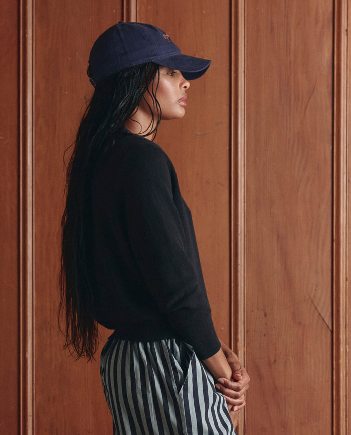 A woman in a navy cap, black top, and striped pants stands in profile against a wooden paneled wall, layering her look with THE GREAT.'s The Shrunken V Neck Cardigan in Black for a modern touch.