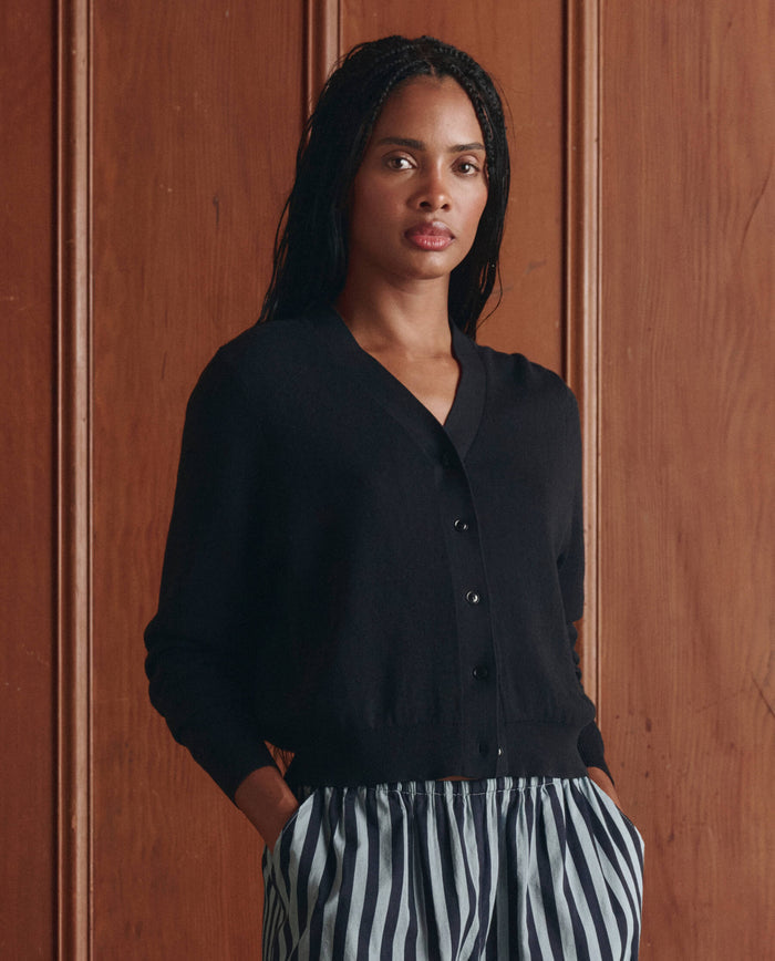 A woman stands in front of a wooden wall, wearing THE GREAT.'s The Shrunken V Neck Cardigan in black and striped pants, with her hands in her pockets.