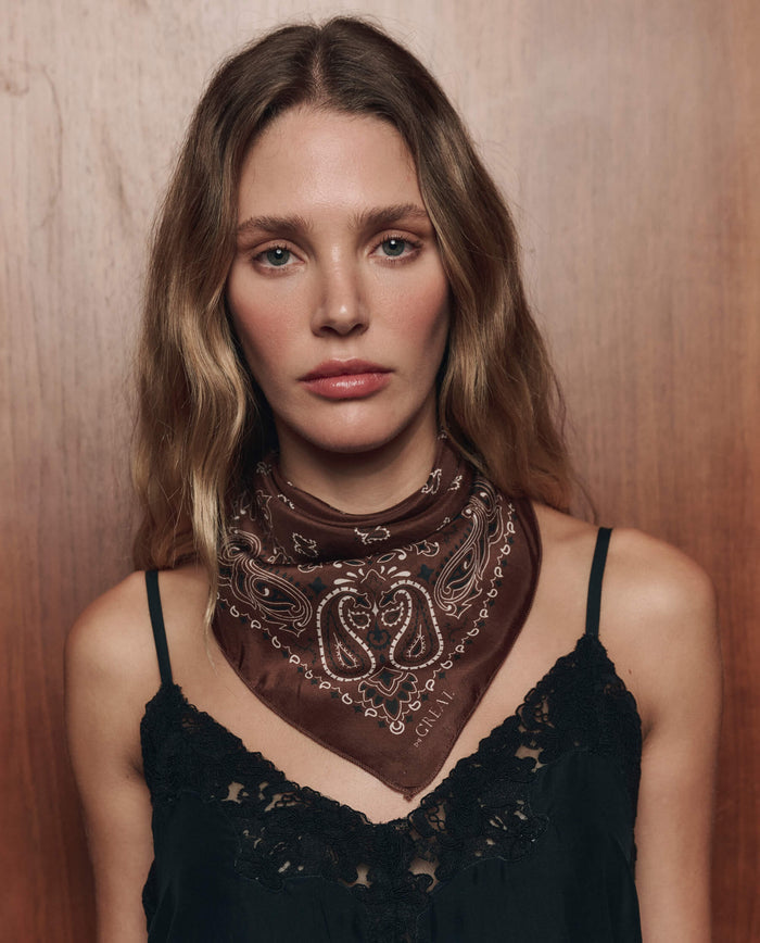A woman with wavy hair stands against a wooden background, wearing a black lace top and The Silk Bandana in Chocolate by THE GREAT. CARRYOVER.