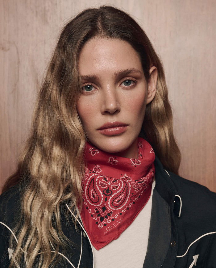 A woman with long, wavy blonde hair wears THE GREAT. CARRYOVER's The Silk Bandana in True Red around her neck, paired with a white shirt and a dark jacket, standing in front of a wooden background.