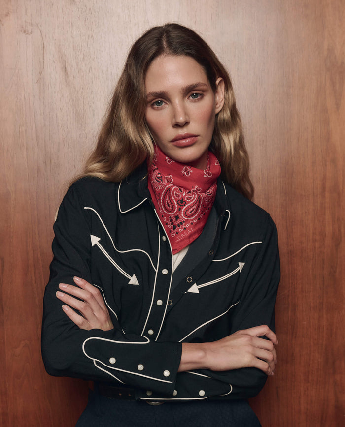 A person with long, wavy hair wears a black western-style shirt featuring white trim and THE GREAT. CARRYOVER's The Silk Bandana in True Red, standing against a wooden background with arms crossed.