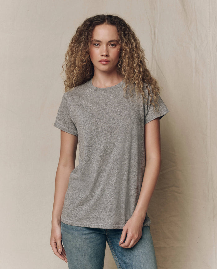A person with curly hair wears THE GREAT. CORE KNITS The Slim Tee. Solid in Heather Grey and blue jeans, standing against a plain, light-colored background.