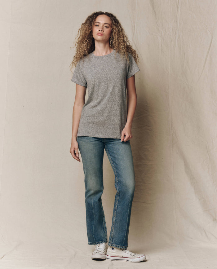 A person with curly hair stands against a beige fabric backdrop, wearing THE GREAT. CORE KNITS' The Slim Tee. Solid in Heather Grey, paired with blue jeans and white sneakers.