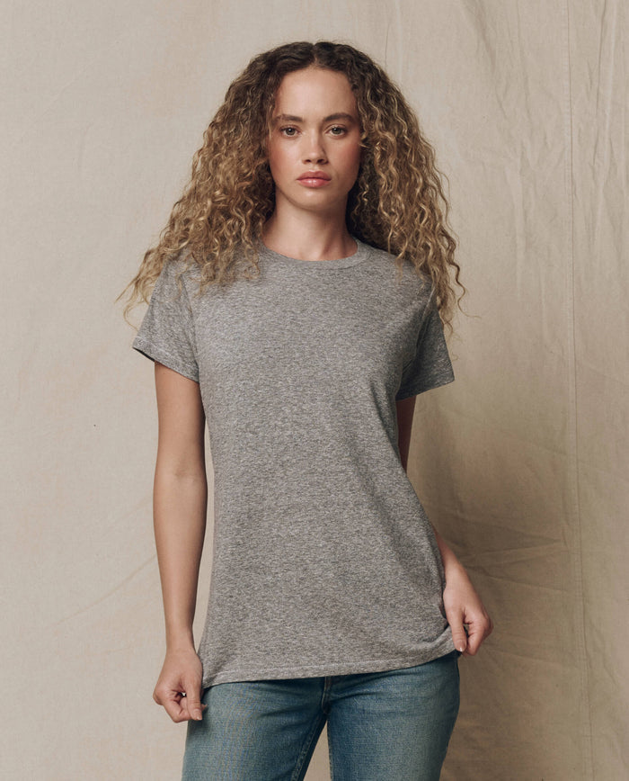 A person with curly hair wears THE GREAT. CORE KNITS The Slim Tee. Solid in Heather Grey and blue jeans, standing against a neutral, textured background.