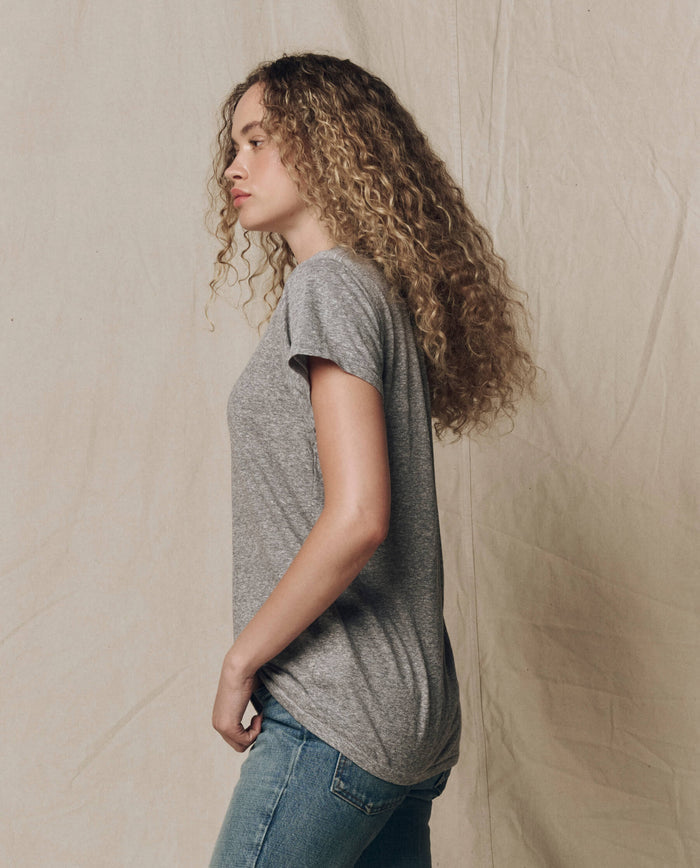 A person with long curly hair stands in profile against a textured backdrop, wearing THE GREAT. CORE KNITS The Slim Tee. Solid in Heather Grey and blue jeans.