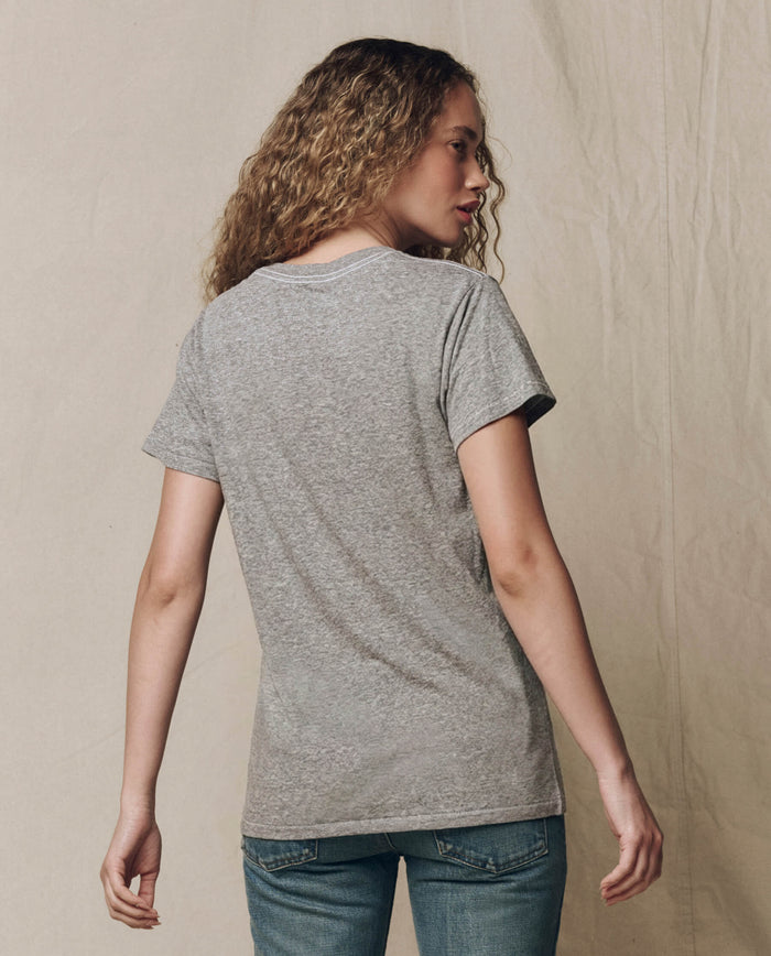 Person with curly hair wears THE GREAT. CORE KNITS The Slim Tee in Heather Grey and blue jeans, standing with their back to the camera against a neutral background.