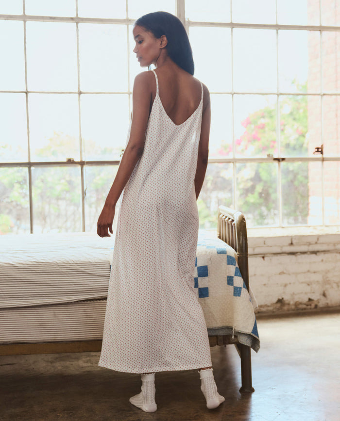 A woman, her back to the camera, stands beside a bed in a bright room wearing THE GREAT. PS26 SLEEP's The Slip Sleeper V Neck Dress in Calico Rose with socks.