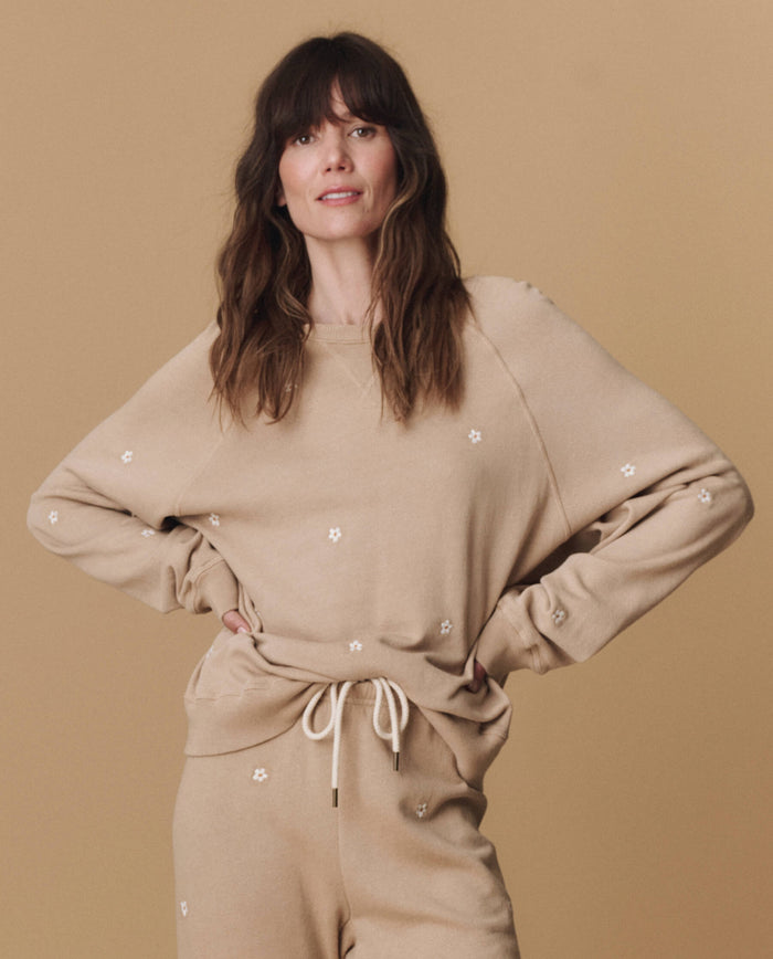 A woman wears THE GREAT. PF25 EMB KNITS The Slouch Sweatshirt in khaki with daisy embroidery, paired with matching pants, standing against a light brown background.