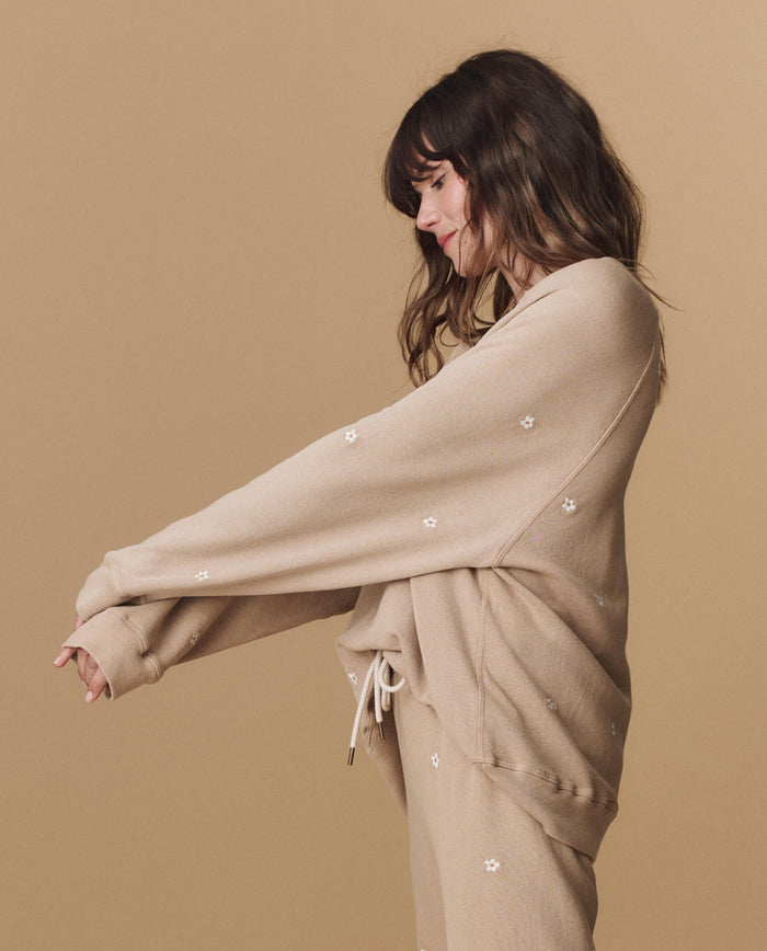 A woman wearing THE GREAT. PF25 EMB KNITS The Slouch Sweatshirt in khaki with daisy embroidery stands against a matching background, facing left with arms extended.