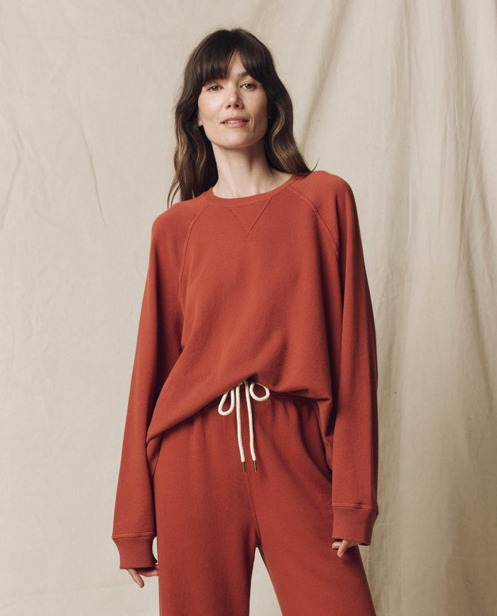 A woman poses against a beige fabric backdrop in THE GREAT. F25 SEASONAL KNITS The Slouch Sweatshirt, Solid -- Brick, paired with matching drawstring pants for a handcrafted Los Angeles look.