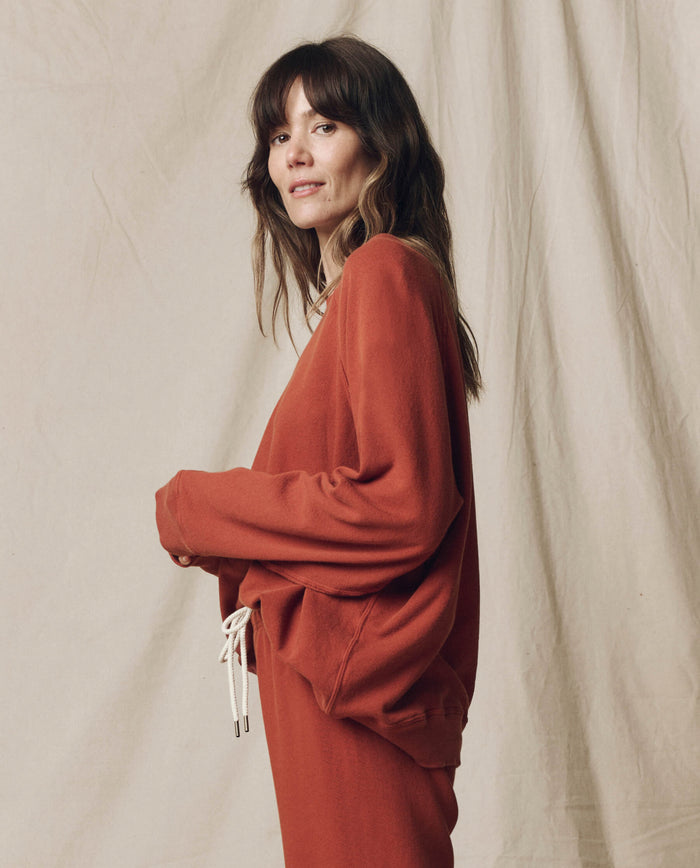 A woman with long brown hair wears THE GREAT.’s The Slouch Sweatshirt in Solid -- Brick from the F25 SEASONAL KNITS, paired with matching pants, standing before a beige fabric backdrop.