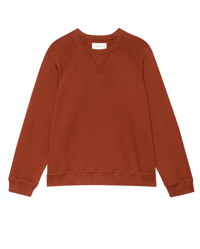 The Slouch Sweatshirt. Solid -- Brick by THE GREAT. F25 SEASONAL KNITS, a rust-colored vintage-inspired piece with long raglan sleeves, ribbed cuffs, and hem, handcrafted in Los Angeles, laid flat on a white background.