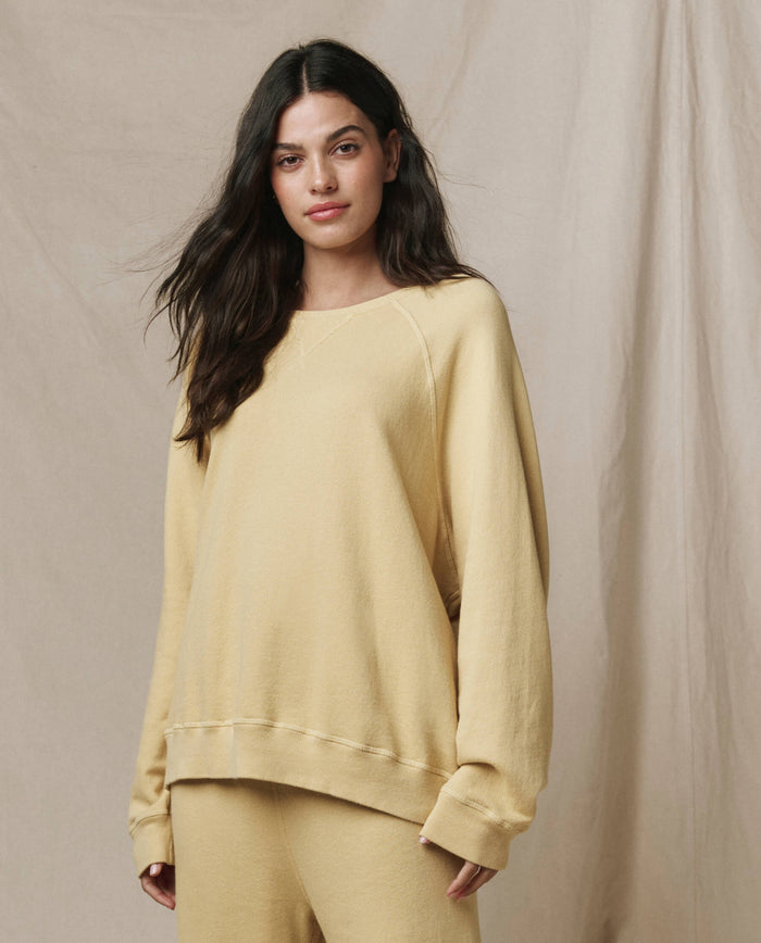 A woman wears The Slouch Sweatshirt in Butter Yellow by THE GREAT. SP26 SEASONAL CORE, paired with matching pants, as she stands before a beige fabric backdrop.