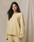 A woman wears The Slouch Sweatshirt in Butter Yellow by THE GREAT. SP26 SEASONAL CORE, paired with matching pants, as she stands before a beige fabric backdrop.