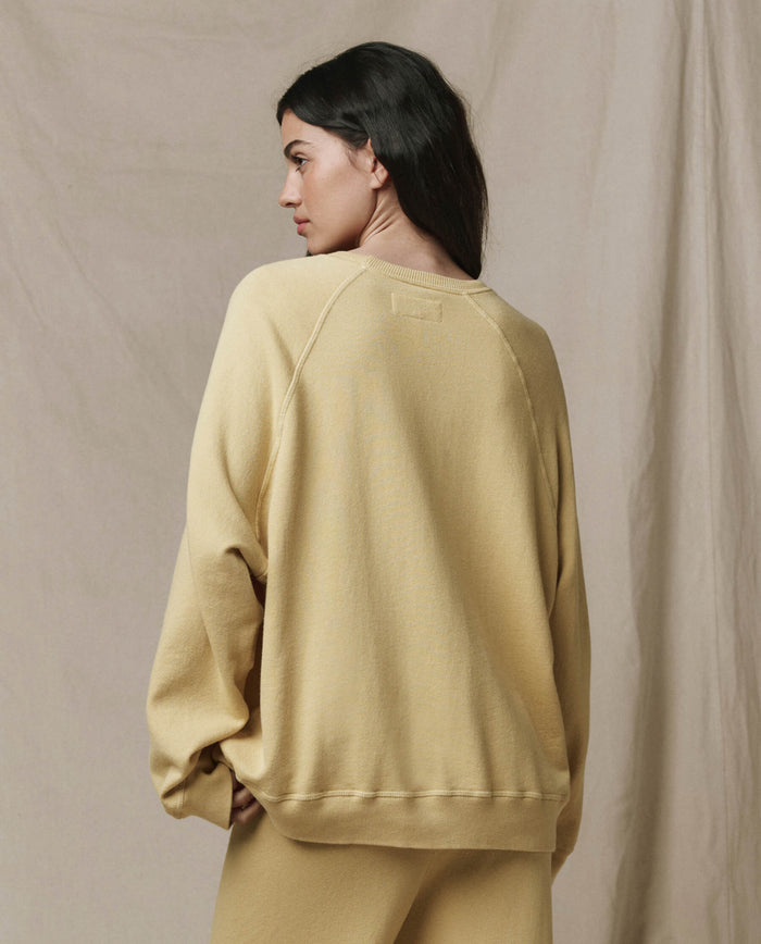 A woman with dark hair, dressed in THE GREAT.'s The Slouch Sweatshirt in Butter Yellow, stands facing away against a neutral fabric backdrop, embodying effortless LA-inspired style from the SP26 SEASONAL CORE collection.