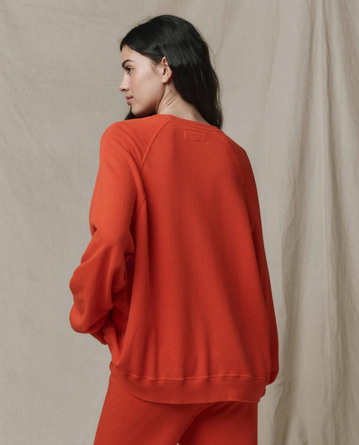 A woman stands sideways, looking left, wearing THE GREAT.'s The Slouch Sweatshirt in Persimmon from the SP26 SEASONAL CORE collection with matching pants against a neutral fabric background.