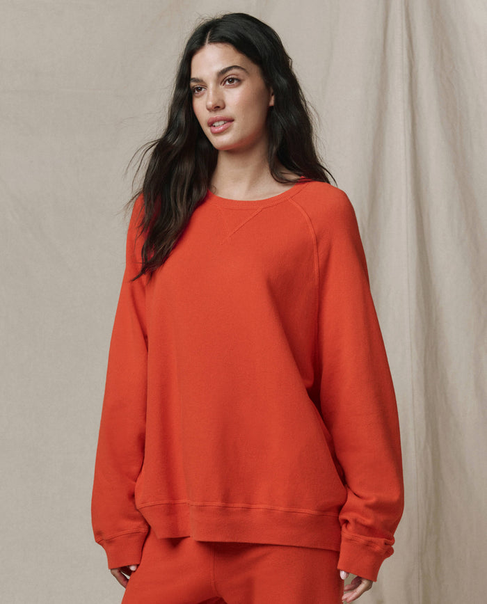 A woman with long dark hair wears THE GREAT. SP26 SEASONAL CORE The Slouch Sweatshirt in Persimmon and matching pants, standing before a neutral fabric backdrop.