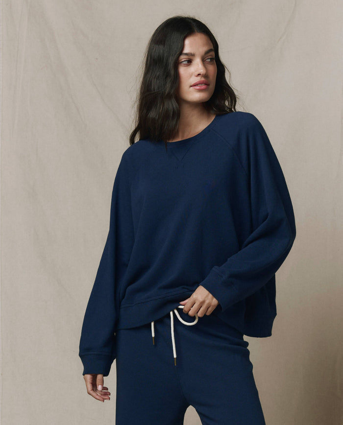 Woman wears THE GREAT. FALL 23 KNITS The Slouch Sweatshirt in True Navy, showing effortless style and handcrafted Los Angeles comfort against a plain beige backdrop.
