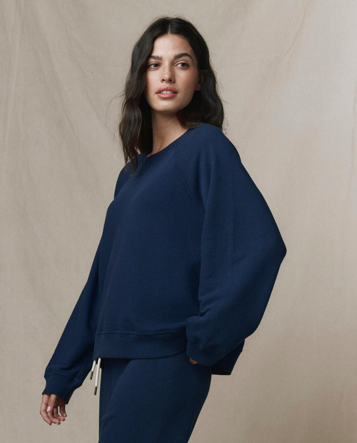 A woman wears THE GREAT.'s The Slouch Sweatshirt in Solid True Navy from the FALL 23 KNITS collection, standing against a neutral beige background.