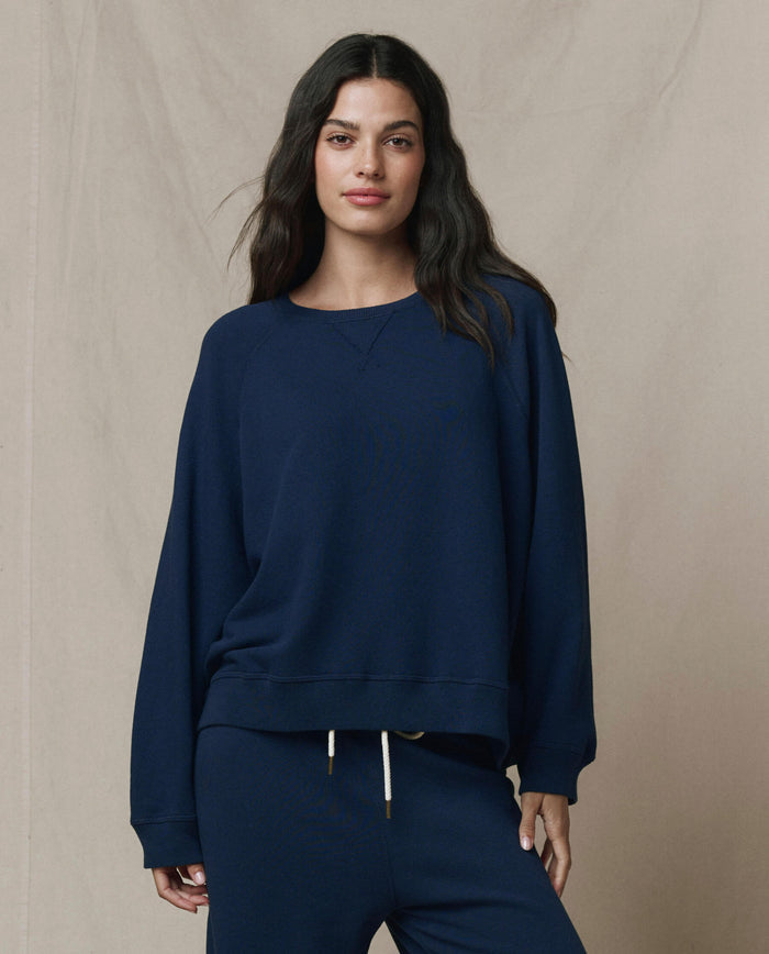 A woman stands against a beige background wearing THE GREAT. FALL 23 KNITS The Slouch Sweatshirt in True Navy and matching sweatpants, handcrafted in Los Angeles, with one hand at her side and the other in her pocket.