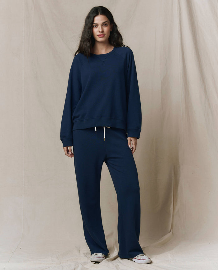 A woman stands facing forward in THE GREAT. FALL 23 KNITS The Slouch Sweatshirt - Solid in True Navy with matching drawstring pants and white sneakers, set against a neutral beige backdrop.