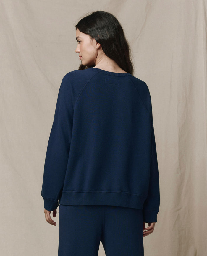 A woman with long dark hair wears THE GREAT.'s FALL 23 KNITS The Slouch Sweatshirt in solid True Navy, paired with matching pants, standing against a beige fabric background and showcasing laid-back Los Angeles style.