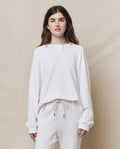A person stands against a beige background wearing The Slouch Sweatshirt in True White by THE GREAT. HOL 24 KNITS, paired with matching pants, giving off a handcrafted vintage vibe, with their hands casually by their sides.