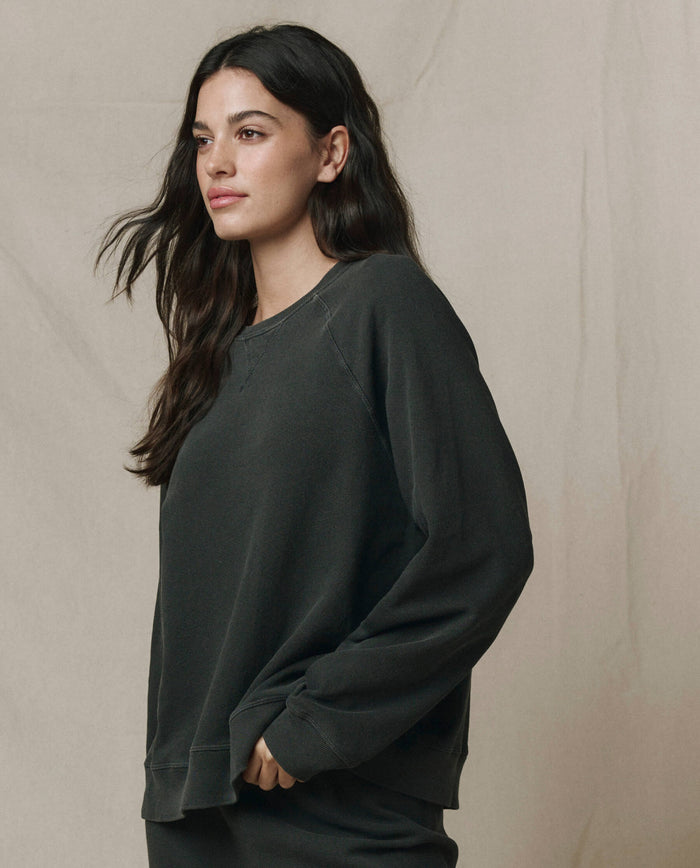 A woman with long dark hair wears THE GREAT. CORE KNITS The Slouch Sweatshirt in Solid -- Washed Black, standing before a neutral beige background.