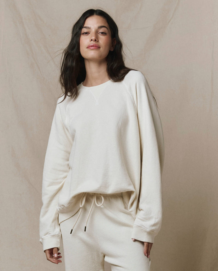 A woman wearing THE GREAT. CORE KNITS The Slouch Sweatshirt in Solid -- Washed White stands in front of a beige backdrop.