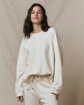 A woman wearing THE GREAT. CORE KNITS The Slouch Sweatshirt in Solid -- Washed White stands in front of a beige backdrop.
