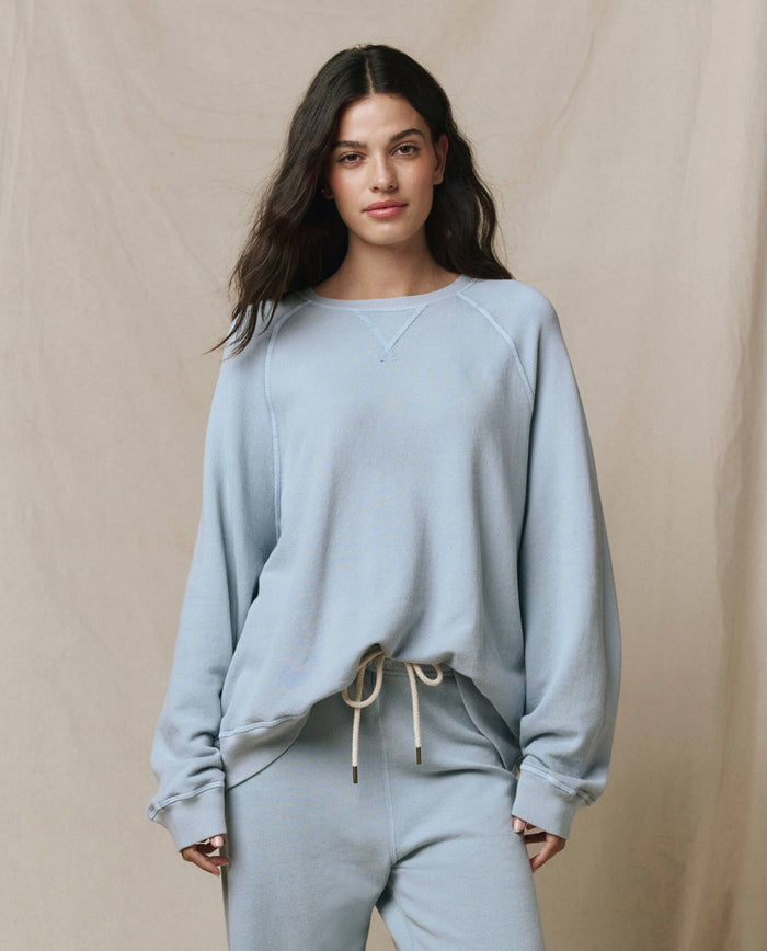 A person wears THE GREAT.'s The Slouch Sweatshirt in Whisper Blue, a vintage-inspired style from the SP26 SEASONAL CORE collection, standing before a neutral fabric backdrop and channeling laid-back Los Angeles fashion.
