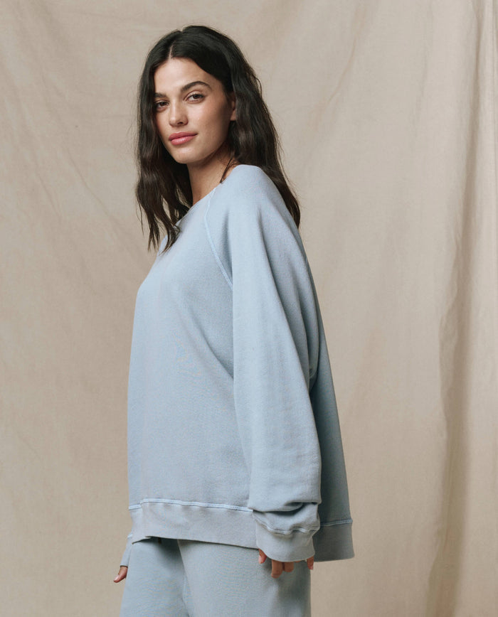 A woman stands sideways against a neutral fabric backdrop wearing THE GREAT.'s The Slouch Sweatshirt in Whisper Blue from the SP26 Seasonal Core collection, paired with matching pants, handcrafted in Los Angeles.