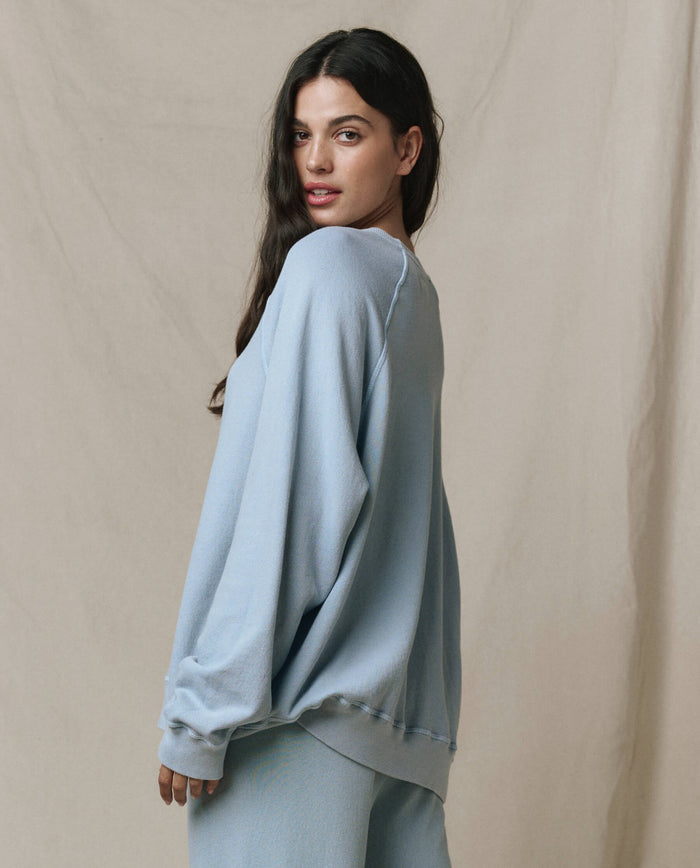 A woman with long dark hair stands sideways against a neutral fabric backdrop, wearing THE GREAT.'s SP26 SEASONAL CORE The Slouch Sweatshirt in Whisper Blue with matching pants.