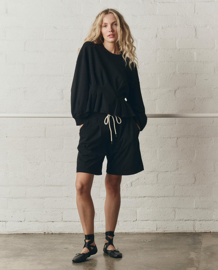 Wearing THE GREAT.'s The Slouchy Corset Sweatshirt in True Black from the SP26 DANCER BODIES collection, a person with long wavy hair stands against a white brick wall in an effortlessly cool Los Angeles-made look.