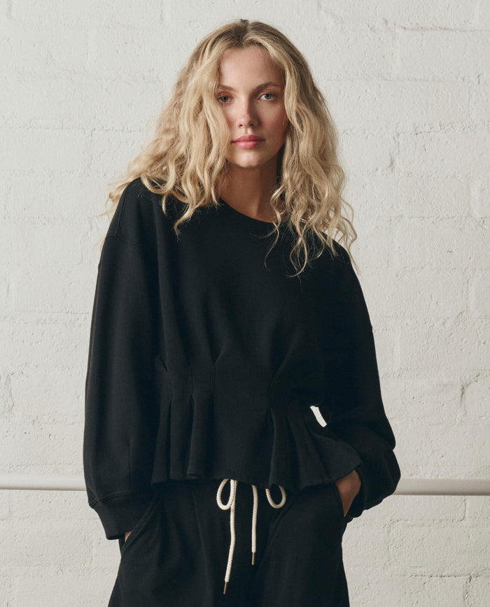 A person with wavy blonde hair wears THE GREAT. SP26 DANCER BODIES The Slouchy Corset Sweatshirt in True Black, paired with black drawstring pants, standing against a white brick wall.
