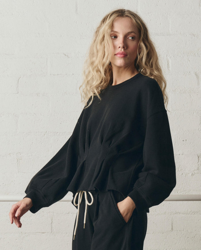 Woman with long, wavy blonde hair wearing THE GREAT. SP26 DANCER BODIES "The Slouchy Corset Sweatshirt" in True Black, handcrafted in Los Angeles, standing against a white brick wall.