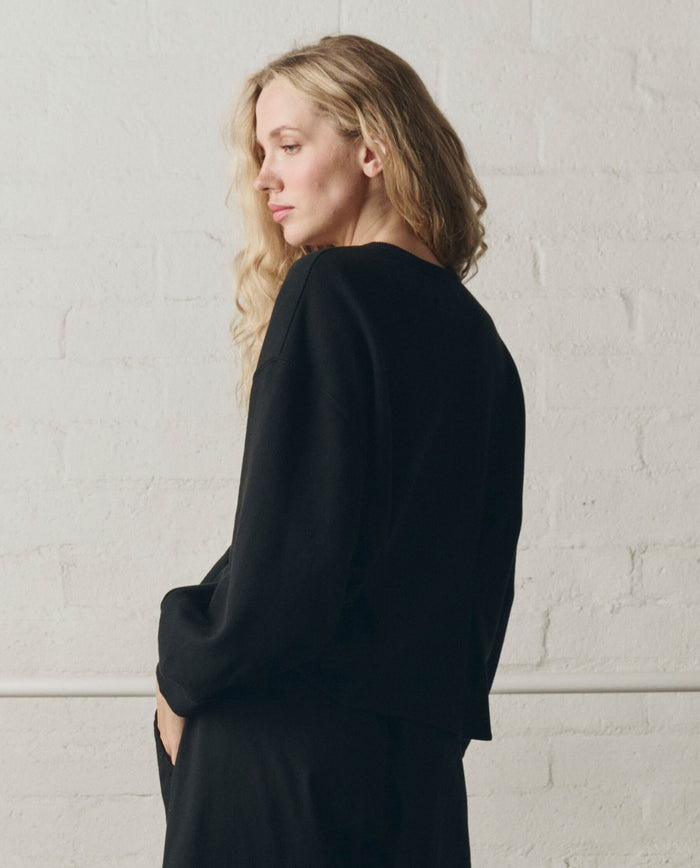 A person with long blonde hair stands sideways against a white brick wall, wearing THE GREAT.'s Slouchy Corset Sweatshirt in True Black from the SP26 DANCER BODIES collection.