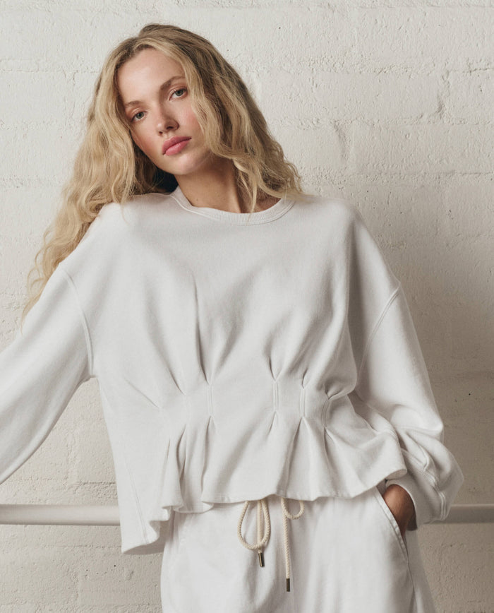 Woman with long blonde hair wears THE GREAT. SP26 DANCER BODIES The Slouchy Corset Sweatshirt in True White, standing by a light brick wall. Handcrafted in Los Angeles for effortless comfort and style.