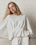 Woman with long blonde hair wears THE GREAT. SP26 DANCER BODIES The Slouchy Corset Sweatshirt in True White, standing by a light brick wall. Handcrafted in Los Angeles for effortless comfort and style.
