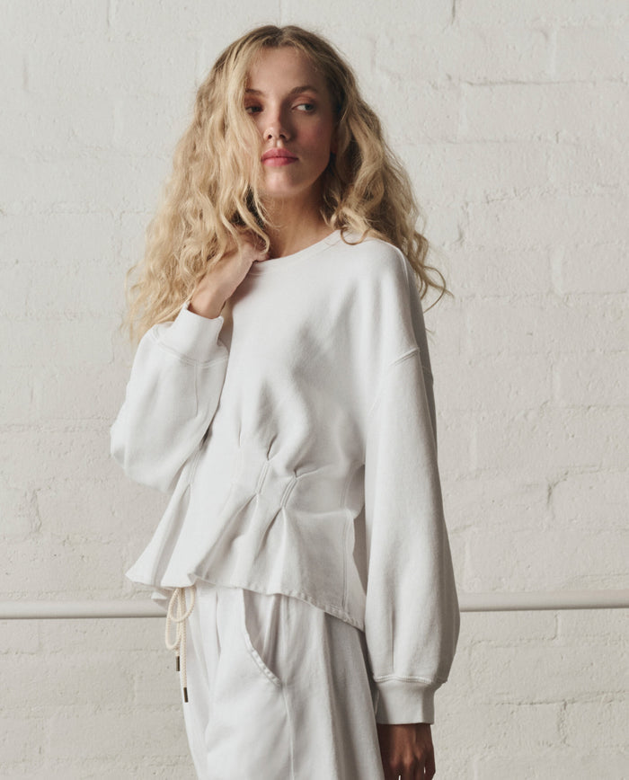 A person with long, wavy blonde hair stands against a white brick wall, wearing THE GREAT.'s Slouchy Corset Sweatshirt in True White from the SP26 DANCER BODIES collection, paired with matching white pants.