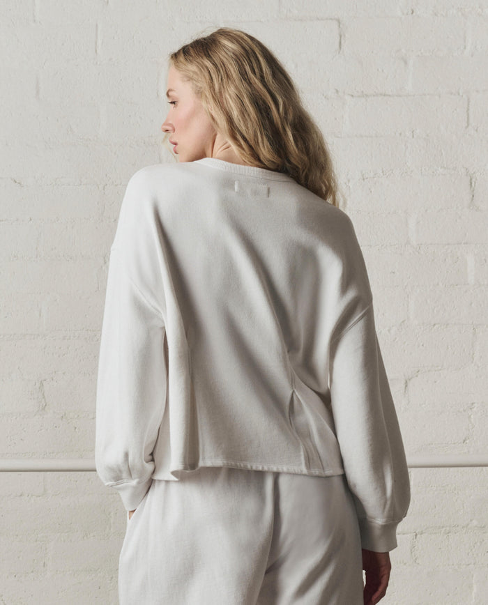A person with long blonde hair wears THE GREAT.'s Slouchy Corset Sweatshirt in True White, part of the SP26 DANCER BODIES collection, standing against a white brick wall and facing away from the camera.