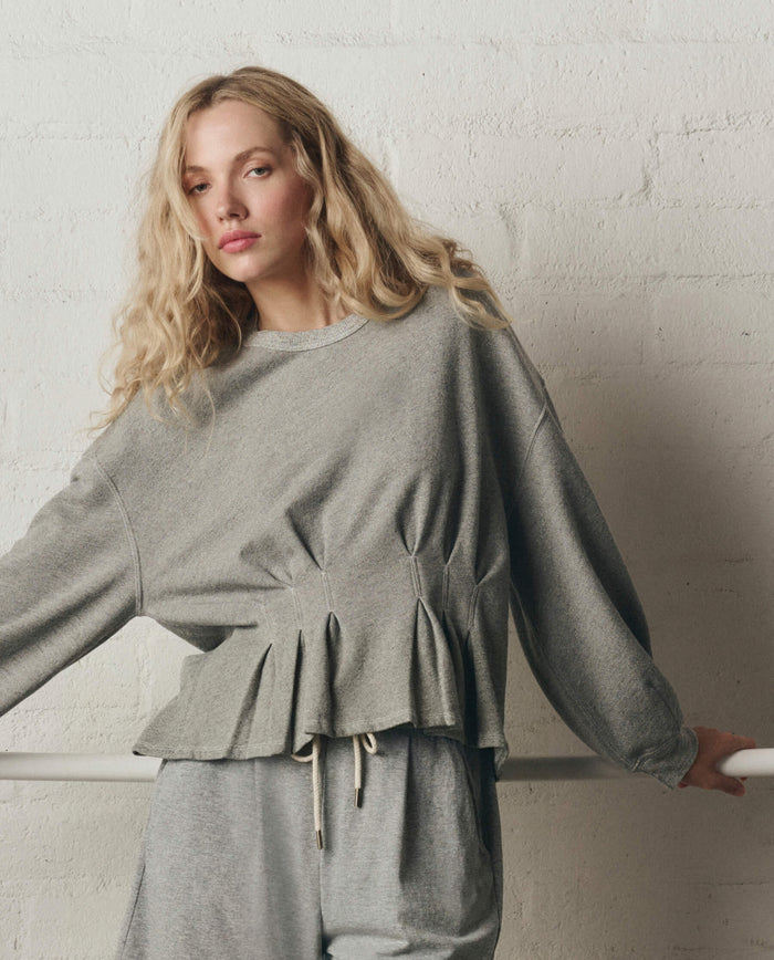 A woman with long blonde hair wears THE GREAT. SP26 DANCER BODIES' The Slouchy Corset Sweatshirt in Varsity Grey, leaning on a white railing against a white brick wall.
