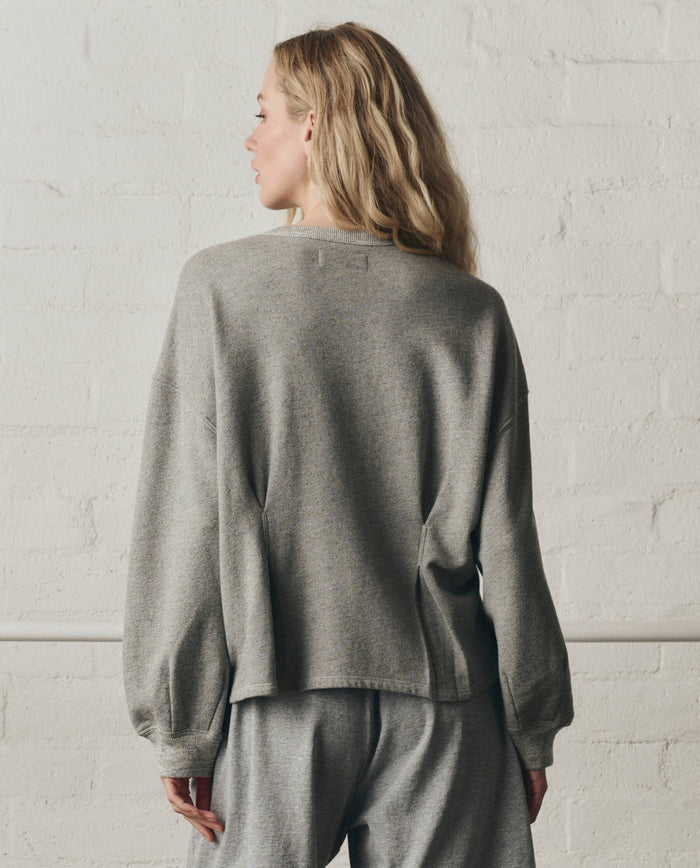 A person with long blonde hair stands with their back to the camera against a white brick wall, wearing THE GREAT. SP26 DANCER BODIES The Slouchy Corset Sweatshirt in Varsity Grey and matching pants.