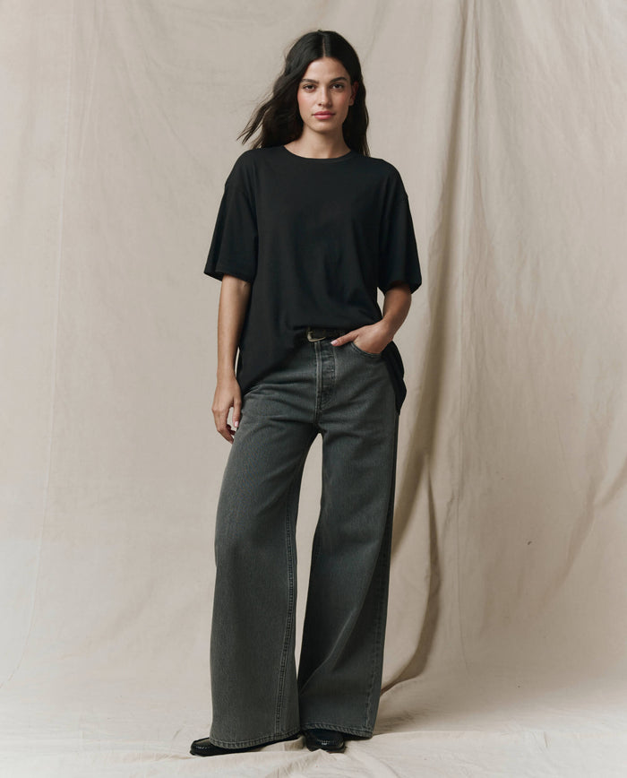 A person stands against a beige fabric backdrop, wearing THE GREAT. SP26 SEASONAL CORE The Slouchy Crew Tee in Almost Black and wide-leg grey jeans, with hands in their pockets.