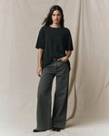 A person stands against a beige fabric backdrop, wearing THE GREAT. SP26 SEASONAL CORE The Slouchy Crew Tee in Almost Black and wide-leg grey jeans, with hands in their pockets.