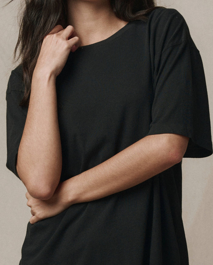 A person with long dark hair wears THE GREAT.'s The Slouchy Crew Tee in Almost Black (SP26 SEASONAL CORE), standing with one arm crossed and the other hand touching their neck.