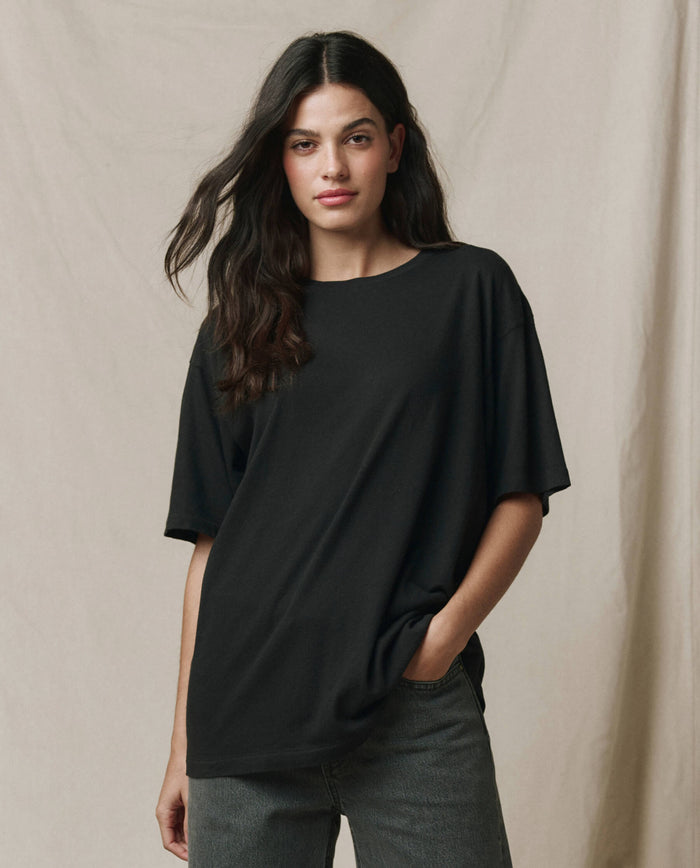 A person with long dark hair wears THE GREAT. SP26 SEASONAL CORE The Slouchy Crew Tee in Almost Black, styled oversized with dark jeans, standing against a neutral fabric backdrop.