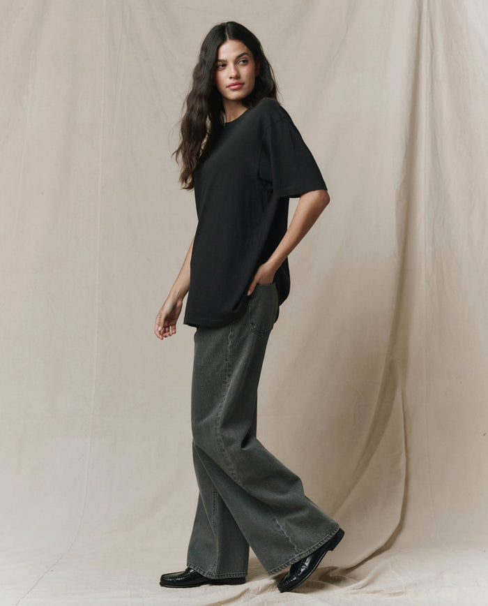 A woman with long dark hair stands before a beige backdrop, wearing THE GREAT. SP26 SEASONAL CORE The Slouchy Crew Tee in Almost Black, paired with gray wide-leg jeans and black shoes.